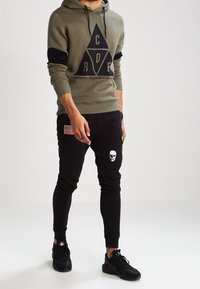 Felpa verde oliva con dettagli neri, caratterizzata da una grafica triangolare nera. Pantaloni da jogging neri con toppe a forma di teschio e bandiera, indossati con sneakers nere.