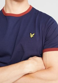 Osoba ubrana w granatowy T-shirt z czerwonym wykończeniem i małym logo żółtego ptaka na piersi, z rękami skrzyżowanymi na tle jasnym.