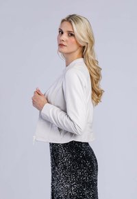 Svetle šedá kožená bomber bunda s přiléhavým siluetem, zapínáním na zip a hladkou texturou. Nosená přes černou sukni posetou flitry.
