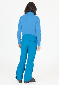 Blå softshell-jakke med høj krave og lynlåslommer, parret med lyseblå bukser med lynlåsdetaljer og en flagrende design.