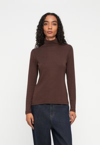 Vrouw met krullend haar draagt een bruine coltrui met lange mouwen en donkerblauwe jeans, staande tegen een effen witte achtergrond.
