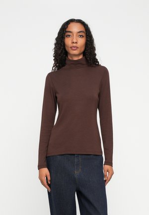 Eine Frau mit lockigem Haar trägt ein braunes Rollkragenlangarmshirt und dunkle blaue Jeans und steht vor einem schlicht weißen Hintergrund.
