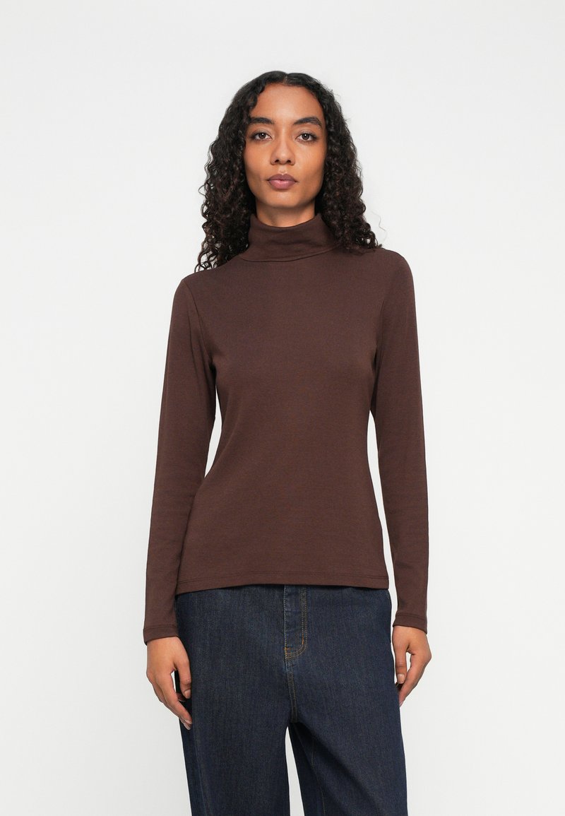Vrouw met krullend haar draagt een bruine coltrui met lange mouwen en donkerblauwe jeans, staande tegen een effen witte achtergrond.
