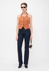 Frau mit einem ärmellosen, orangefarbenen, knopfgeschlossenen Halstopp, dunkelblauen High-Waist-Jeans, schwarzen spitzen Schuhen, Sonnenbrille und einer Tasche im Leopardenmuster.