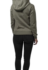 Sudadera con capucha verde oliva de corte holgado, con puños y dobladillo acanalados, llevada sobre unos vaqueros ajustados negros, vista desde atrás.