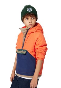 Veste à capuche orange avec des accents bleu marine, dotée d'une poche avant et du logo Timberland. Bonnet en laine vert avec un détail de logo.