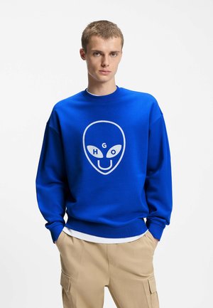 Junger Mann trägt ein knallblaues Sweatshirt mit einem weißen Alien-Gesicht und den Buchstaben H, G, O auf der Vorderseite, kombiniert mit beigen Cargohosen.