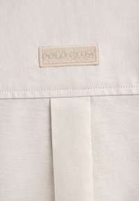 Torba płócienna w jasnym beżu z teksturowaną powierzchnią, z logo naszytym na metce oraz klapką z detalem w postaci zakładki.