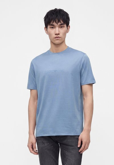 Νέος άνδρας που φοράει ένα ανοιχτό γαλάζιο t-shirt Emporio Armani και μαύρα τζιν, στέκεται μπροστά σε λευκό φόντο.