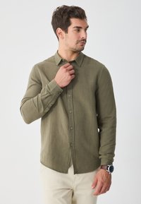 Olivgrüne Knopf-up-Bluse mit Kragen, langen Ärmeln, grünen Knöpfen und geradem Saum. Der Stoff wirkt strukturiert und leicht.