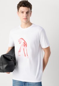 Fehér pamut póló piros oroszlán grafikával. Kerek nyak, rövid ujjú, laza szabású. A modell bal kezében fekete táska van.