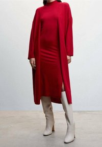 Femme portant une robe midi rouge à manches longues avec une fente sur la cuisse, associée à un long cardigan rouge assorti, portée avec des bottes blanches à talons hautes jusqu'aux genoux.