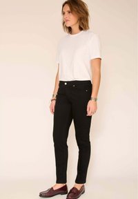 Femme debout, portant une chemise blanche à manches courtes, un pantalon noir ajusté, des mocassins marron foncé et des bracelets argentés aux deux poignets.