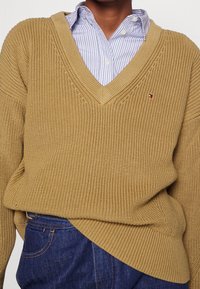 Strickpullover in Senfgelb mit tiefem V-Ausschnitt, strukturiertem Muster und gerippten Bündchen. Über einem hellblauen, gestreiften Hemd in Weiß getragen.