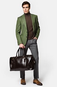 Zielona marynarka w kratę, brązowy golf, szare spodnie i brązowe skórzane buty. Trzyma ciemnobrązową skórzaną torbę typu duffel.