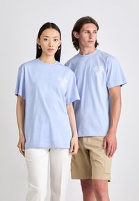 Zwei Personen tragen hellblaue, kurzärmlige T-Shirts mit Rundhalsausschnitt und einem Blumenmuster auf der Brust. Der Stoff scheint weich und glatt zu sein.