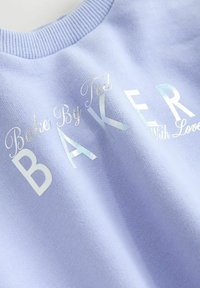 Tessuto di un leggero viola con scritte argentate brillanti "Baker" e parole corsive "Baked By Ted" e "With Love" su un indumento lavorato a maglia.