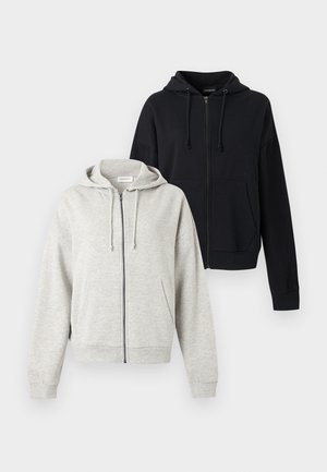 Jeune femme debout en tenue décontractée : sweat à capuche gris zippé, débardeur noir, short en jean gris, et baskets noires à rayures blanches.
