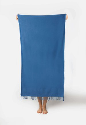 Henkilö seisoo paljain jaloin pitäen suurta sinistä kangasta pystyasennossa, peittäen kokonaan ylävartalon ja kasvot, yksinkertaisen valkoisen taustan edessä.