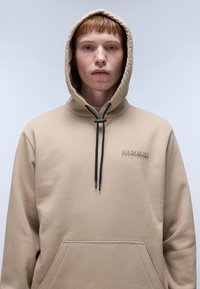 Sudadera con capucha beige con cordones negros, bolsillo frontal y detalle del logo en el lado izquierdo del pecho; textura de tela suave.