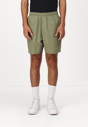 Homme portant un short taille élastique vert olive, des chaussettes blanches et des baskets blanches sur un fond blanc uni.