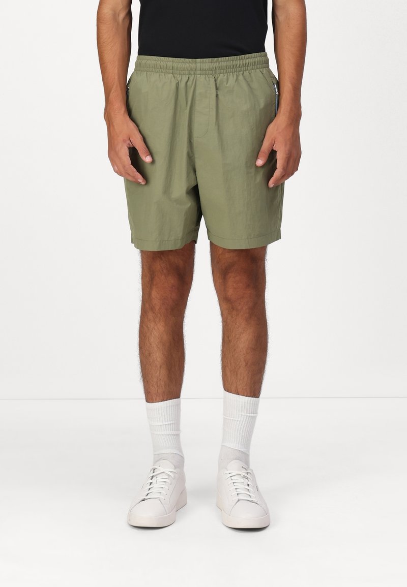 Homme portant un short taille élastique vert olive, des chaussettes blanches et des baskets blanches sur un fond blanc uni.