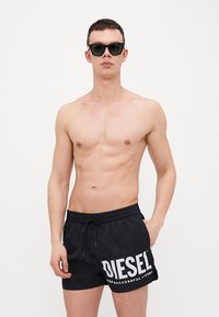 Shorts de bain noirs avec un cordon de taille et un logo "DIESEL" blanc, portés par un mannequin masculin torse nu, associés à des lunettes de soleil noires.
