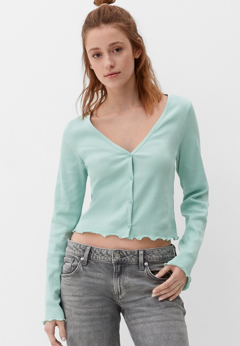 Mintgroene geribde cardigan met lange mouwen, een V-halslijn en golvende randjes. Voor te sluiten met vijf knopen aan de voorkant. Draagt met grijze jeans.