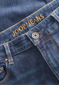 Mörkblå denimjeans har en gyllene broderad varumärkesnamn "JOOP!JEANS" på midjebandet, silverfärgad knappstängning och synliga sömnadsdetaljer.
