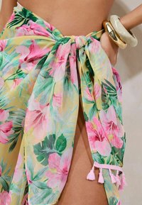 Kimonoflora keltamarang kuning, dengan bunga hibiskus merah jambu dan daun hijau, diikat di pinggang dengan aksen tassel merah jambu di ujungnya.
