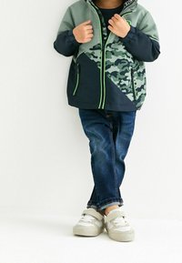 Grön och marinblå färgblockerad jacka med kamouflage-mönster, dragkedja och neonlysande detaljer, parat med mörka jeans och ljusa sneakers.