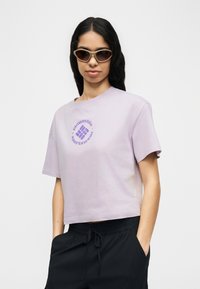 Γυναίκα που φοράει cropped μπλουζάκι Columbia Sportswear σε ανοιχτό μωβ χρώμα, μαύρο παντελόνι με κορδόνι και μπεζ γυαλιά ηλίου σε σχήμα cat-eye, στέκεται με τα χέρια στις τσέπες.