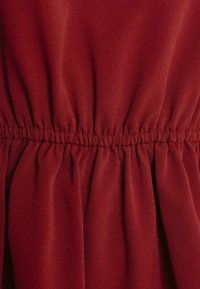 Červená tkanina s jemně strukturovaným povrchem, s nabíranými detaily v pase, které vytvářejí řasený efekt napříč oděvem.