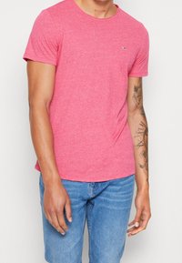 Red short-sleeve t-shirt made of soft fabric with a crew neck. Features a small logo on the chest and a relaxed fit. Worn with blue denim jeans.