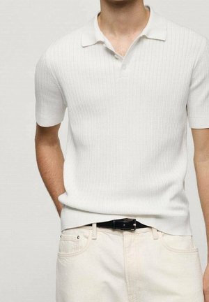 Mann trägt ein eng anliegendes weißes, geripptes Poloshirt mit kurzen Ärmeln, in cremefarbene Hose mit schwarzem Gürtel gesteckt, Hand in der Tasche.