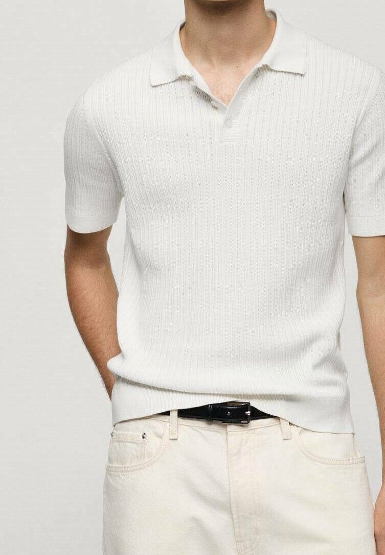 Homme portant un polo blanc ajusté côtelé à manches courtes, rentré dans un pantalon écru avec une ceinture noire, main dans la poche.