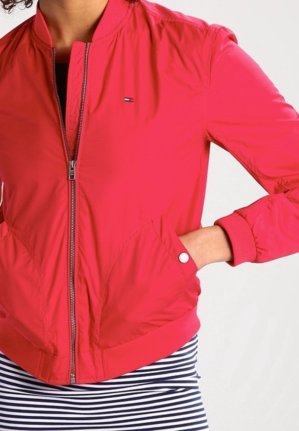 Personne portant une veste zippée rouge vif, une main dans la poche et une jupe rayée noir et blanc en dessous.
