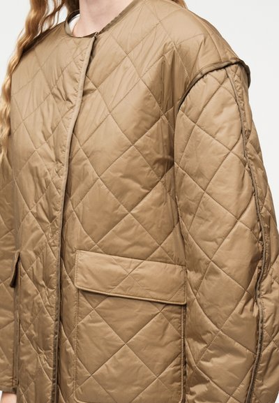 Κοντινό πλάνο ενός ατόμου που φοράει μπεζ μπουφάν με ραφές σε σχήμα ρόμβου και μεγάλες μπροστινές τσέπες με καπάκι.