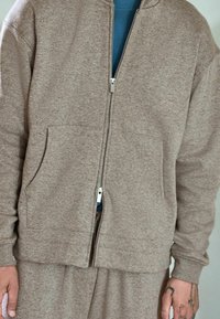 Brun hættetrøje med lynlås lavet af tekstureret stof, der har to forlommer og en ribbet kant, lagdelt over en blå skjorte.