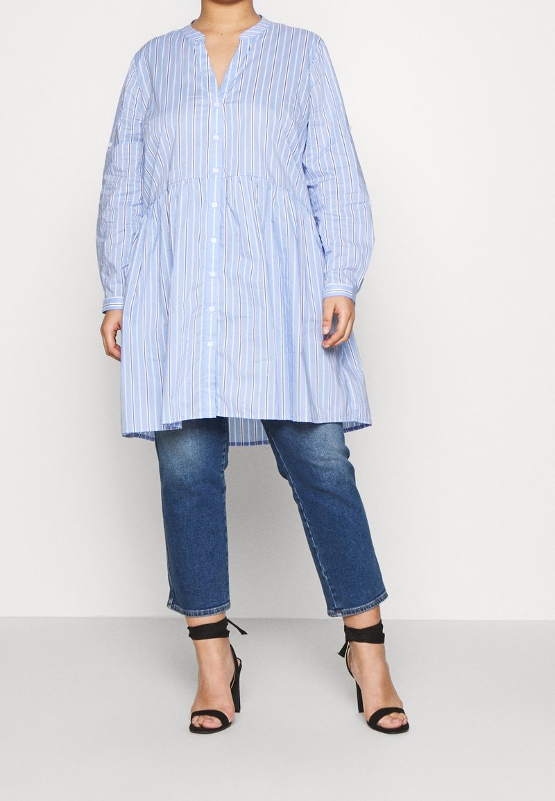 Blauwe en witte gestreepte lange blouse met een v-hals, knoopsluiting aan de voorkant en een geraffineerde taille, gecombineerd met denim jeans en zwarte hoge hakken met bandjes.