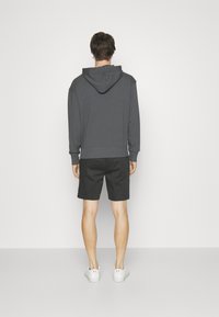 Felpa grigia con tasca a marsupio e cappuccio con cordino, abbinata a pantaloni corti neri texturizzati e scarpe da ginnastica bianche, vista da dietro.
