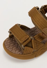 Brun sandal med textilremmar, texturerad gummisula, justerbara kardborrestängningar och en mönstrad fotbädd för komfort.