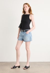 Svart ermeløs topp, lyseblå mønstrete denimshorts, svart belte og svarte hæl-sandaler, stående på en beige gulv.