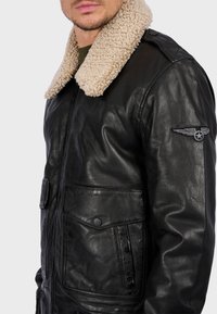 Černá kožená bunda s béžovým umělým beránkem na límci, se zipem a dvěma předními kapsami; na rukávu je vyšitý emblém s křídly.