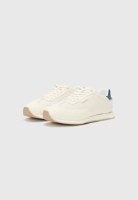 White athletic sneakers with mesh and smooth synthetic material, rounded toe, beige sole, and navy blue heel accents.