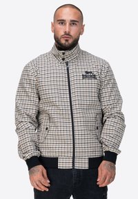 Kurtka w kratę w beżowych i brązowych odcieniach, z zamkiem z przodu, wysokim kołnierzem i prążkowanymi mankietami. Posiada boczne kieszenie i logo na piersi.