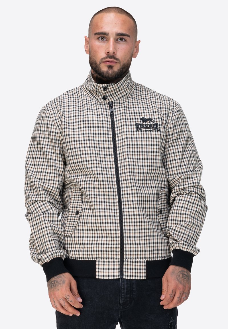 Kurtka w kratę w beżowych i brązowych odcieniach, z zamkiem z przodu, wysokim kołnierzem i prążkowanymi mankietami. Posiada boczne kieszenie i logo na piersi.