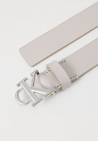 Calvin Klein világosszürke bőr öv ezüst fém csattal, amelyen a "CK" logó található, sima felületű, és minimális varrási részletekkel rendelkezik.