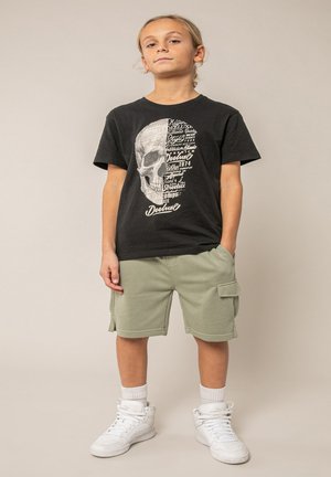 Enfant debout, les mains dans les poches, portant un T-shirt noir avec un motif de crâne, un short cargo vert clair, des chaussettes blanches et des baskets blanches.