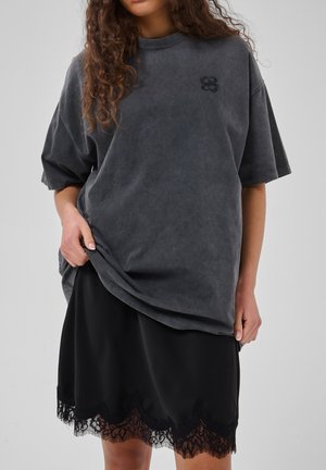 Kobieta w za dużym, spranym czarnym t-shircie z małym logo na piersi oraz w czarnej spódnicy z koronkową lamówką przy dole, stoi swobodnie.